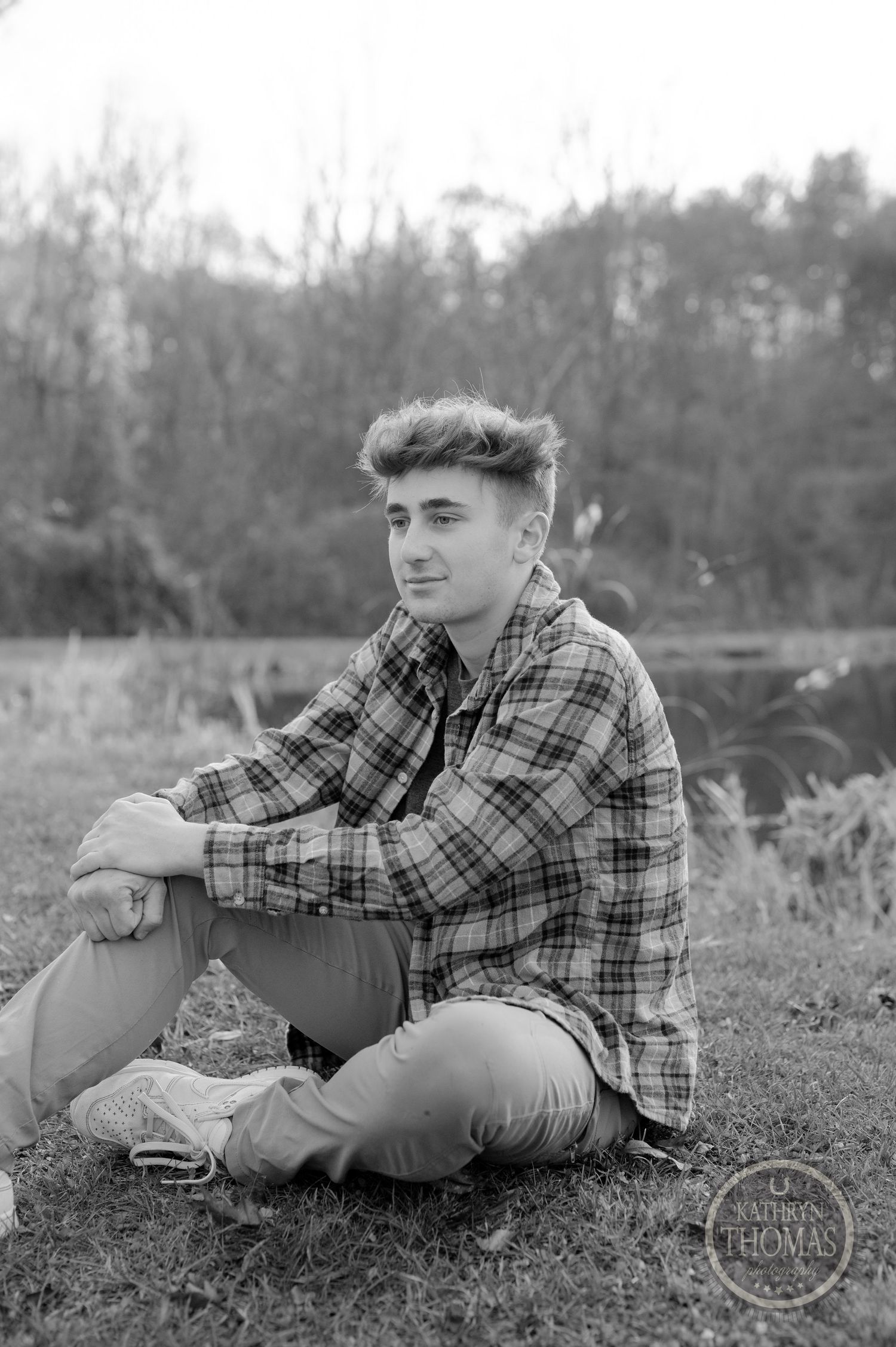 Black and white portrait of someone sitting on grass wearing plaid and casual attire.