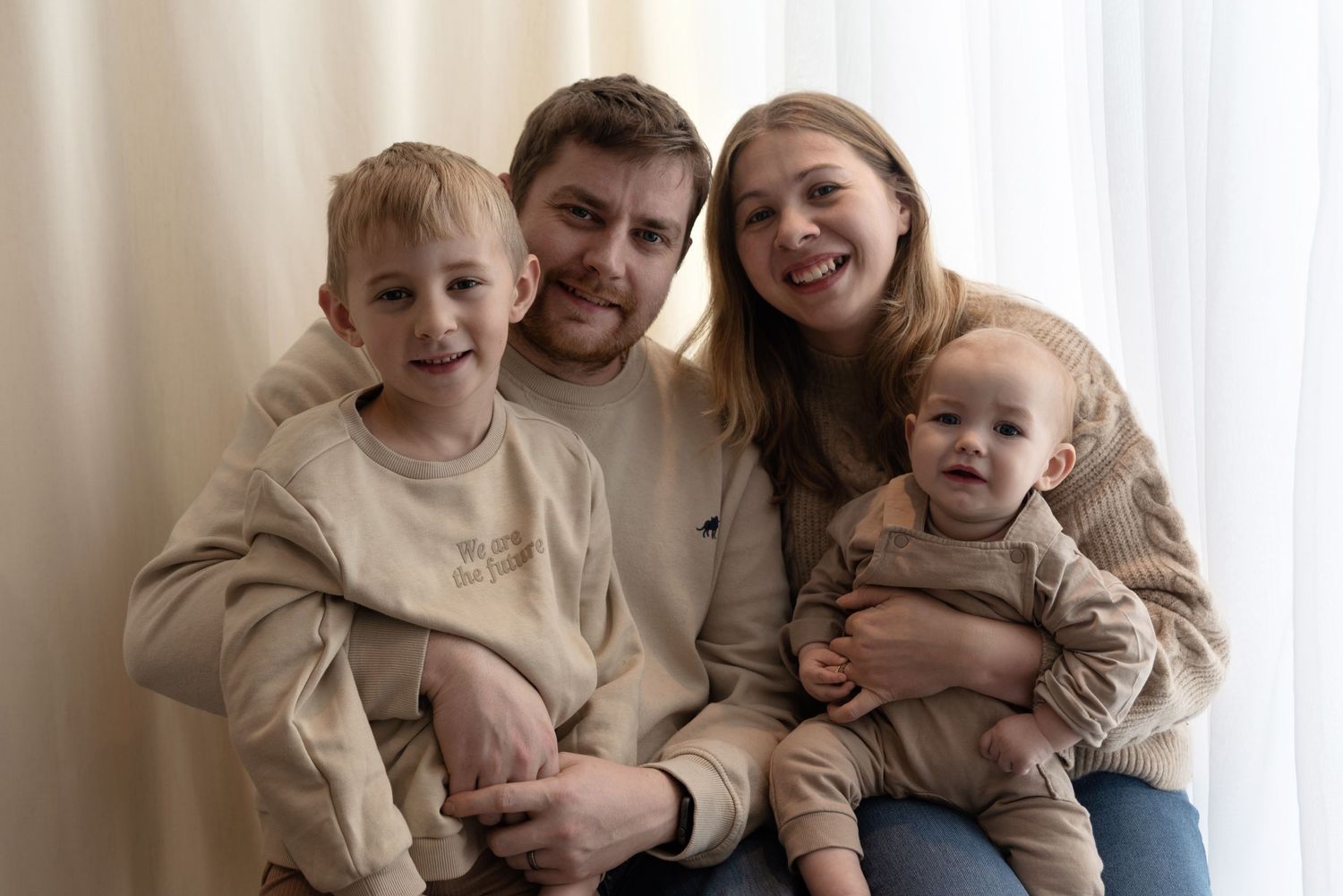 A family of four wearing beige sweaters sits together against a white curtain backdrop, sharing warm smiles.
