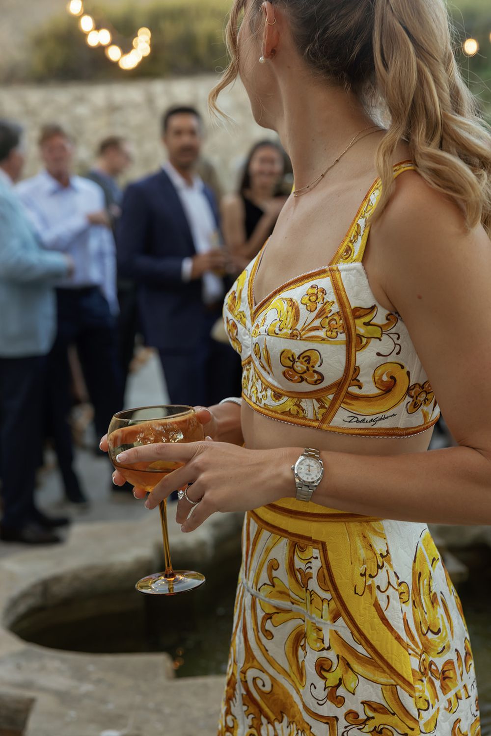 A guest in a yellow patterned dress holds a champagne glass at an outdoor event.