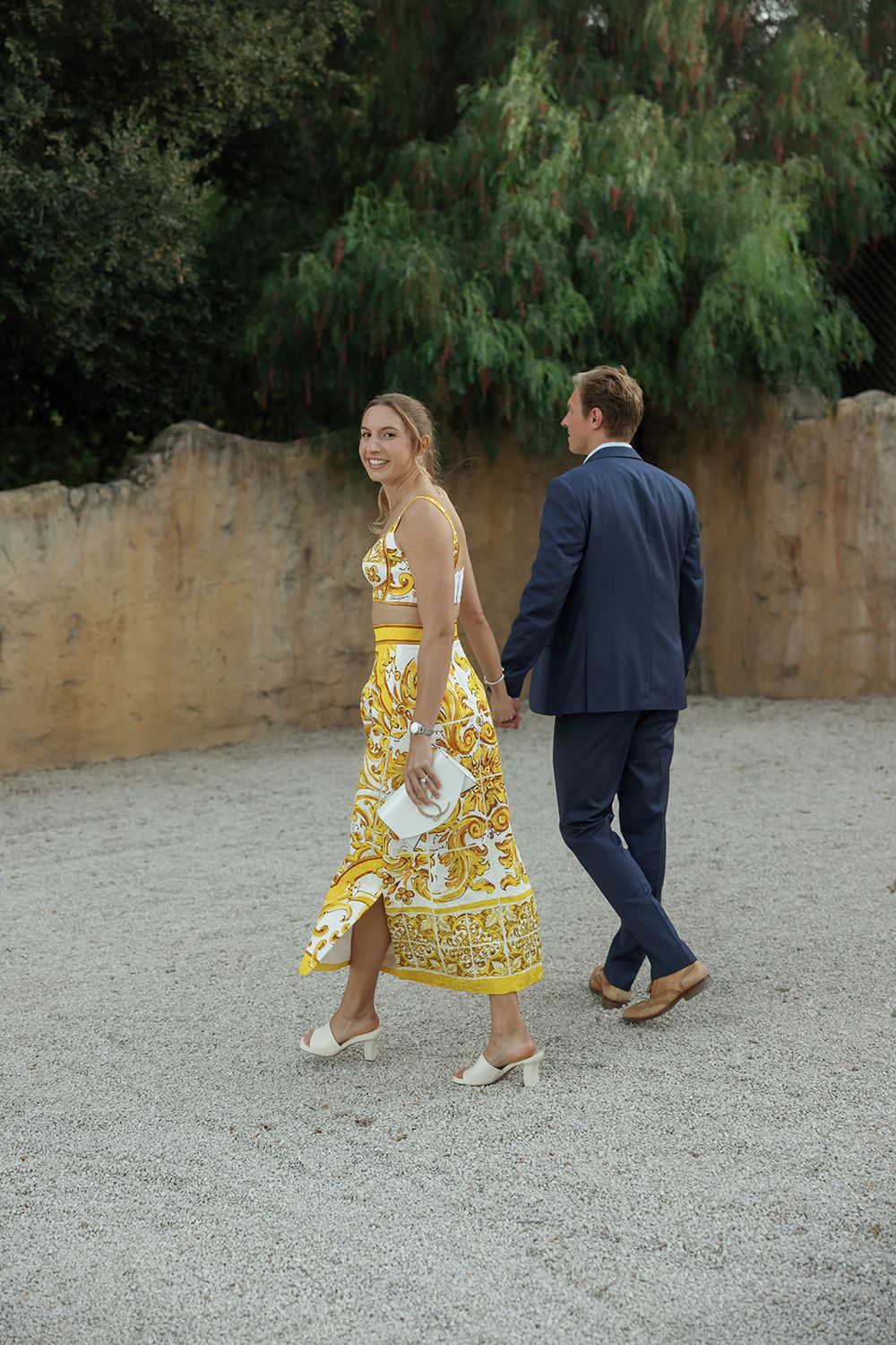 A couple walking on a gravel path, with one wearing a yellow patterned summer dress.