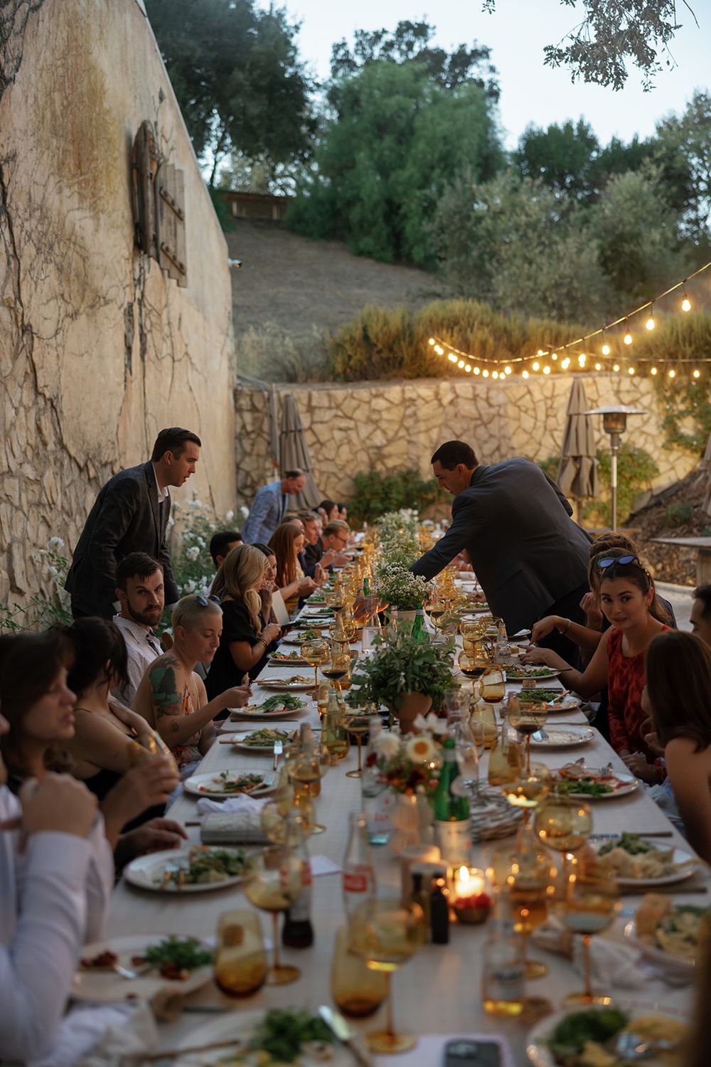 Guests enjoy an elegant outdoor dinner party under string lights with a rustic stone wall backdrop.