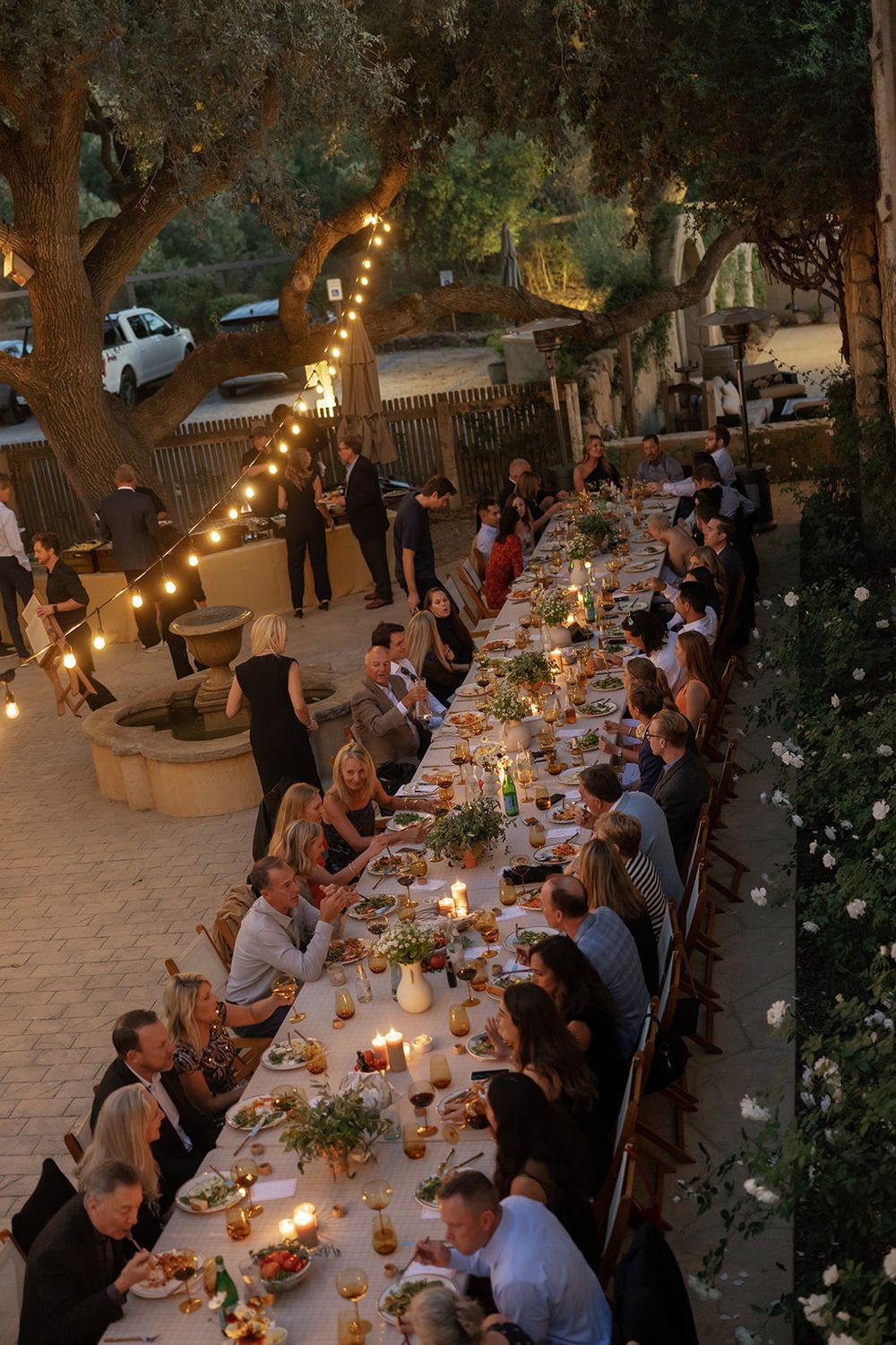 Long table dinner party set in a garden courtyard with string lights overhead at dusk.