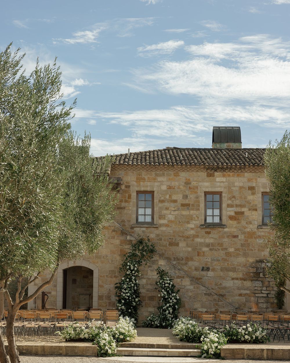 A rustic stone building with olive trees and white floral landscaping creates a Mediterranean atmosphere.
