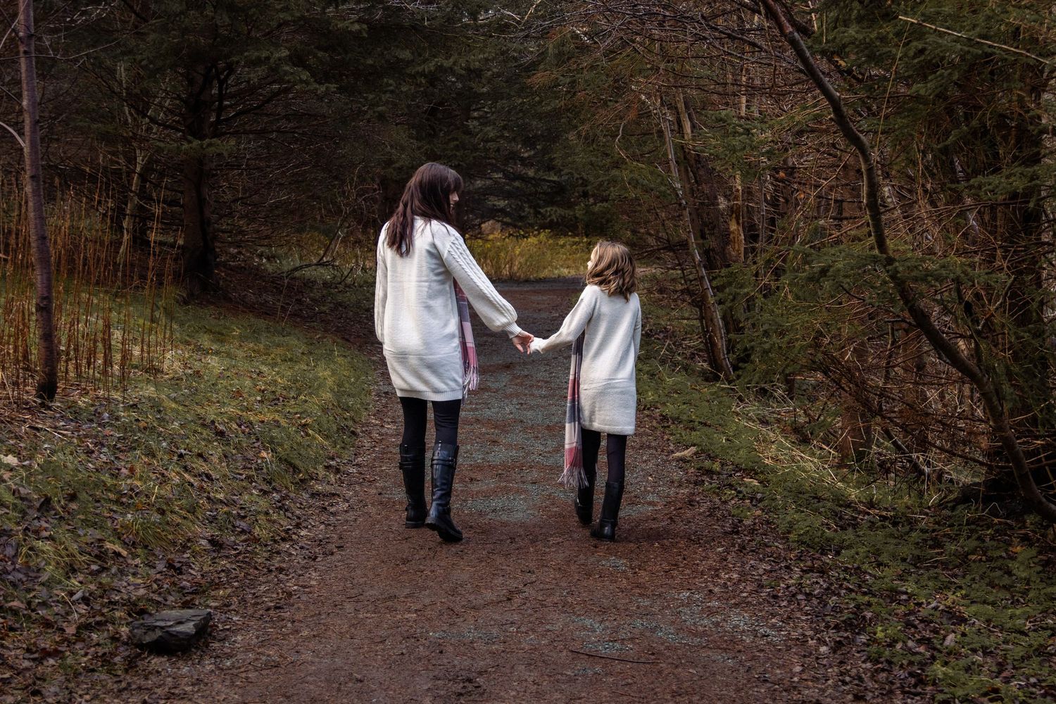 Mom and daughter in white sweaters holding hands while walking on a brown dirt path through autumn woods.