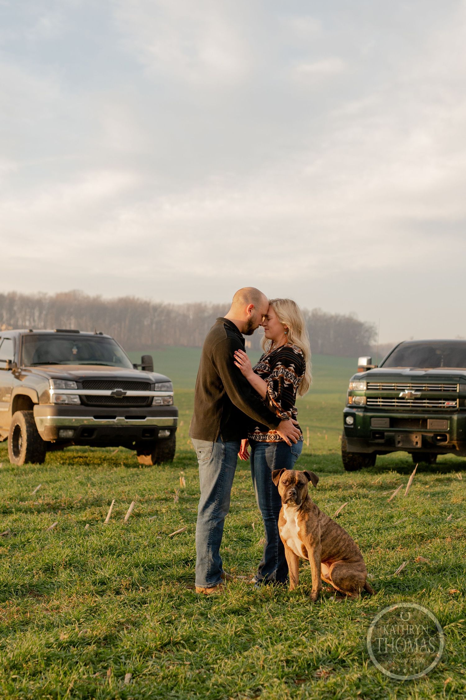 A couple poses with their pit bull dog between two pickup trucks in a grassy field.