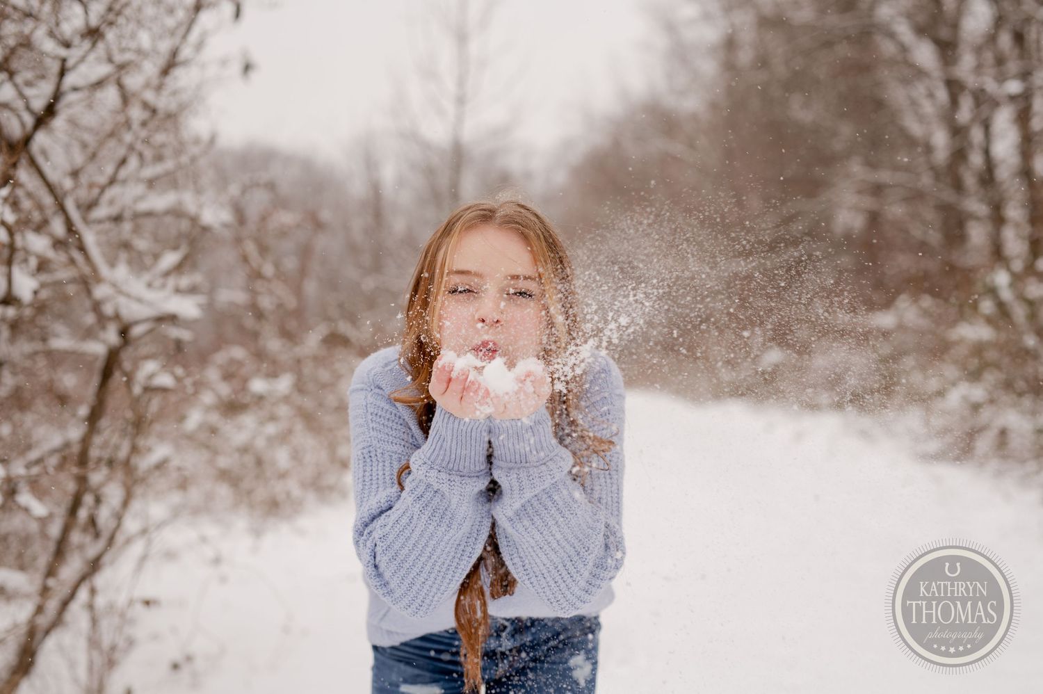 Someone playfully blows snowflakes while wearing a blue sweater in a wintry forest setting.