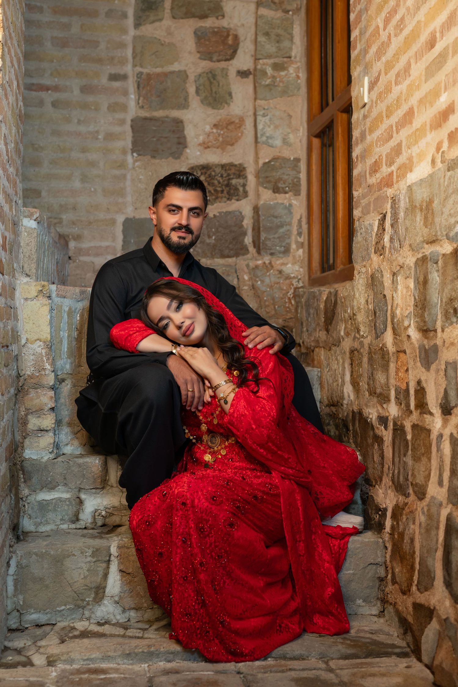 A couple sits together on stone steps against a rustic wall backdrop, with the woman wearing an elegant red lace gown.