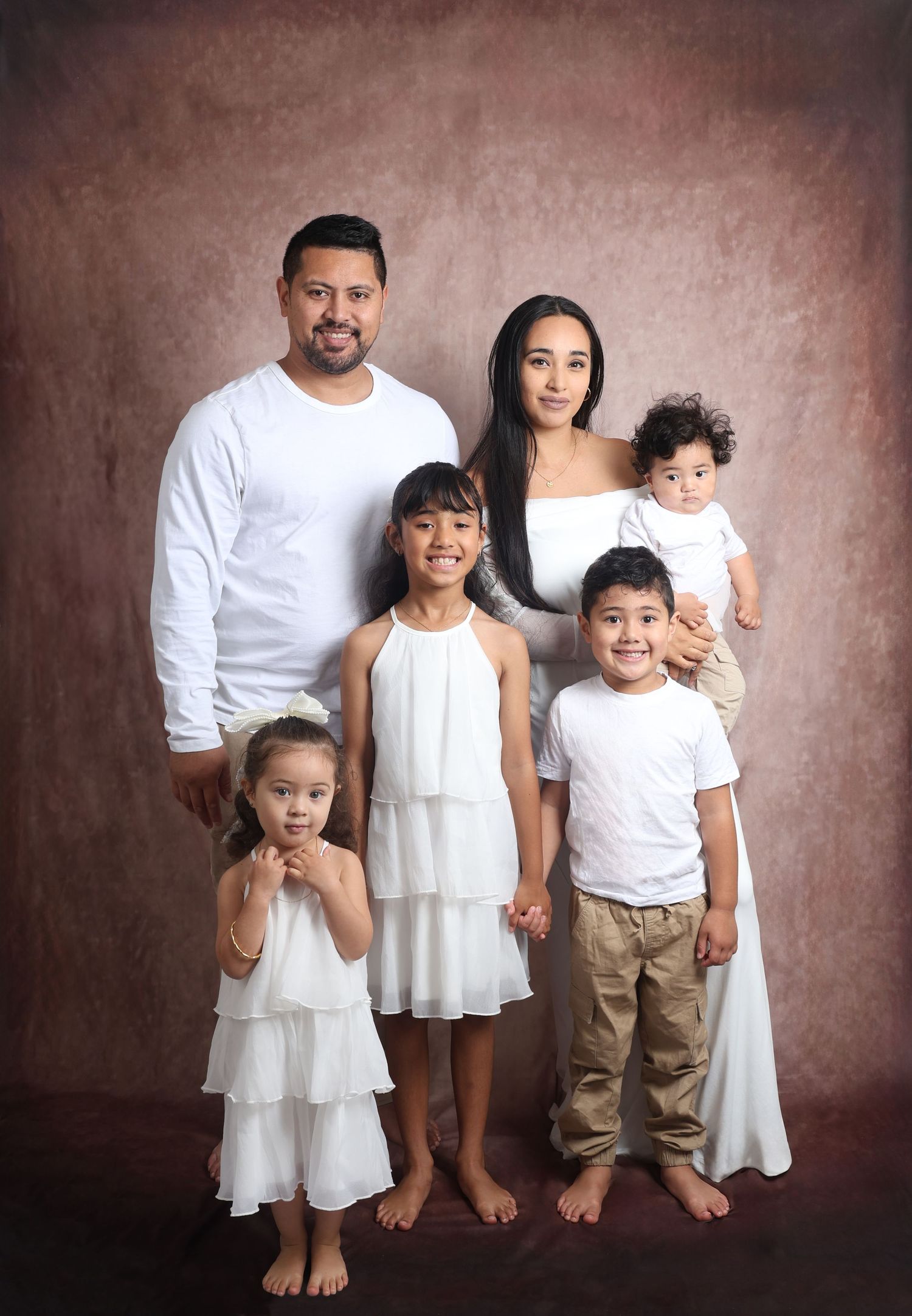 Studio family portrait series with coordinating white and beige outfits against brown backdrop.