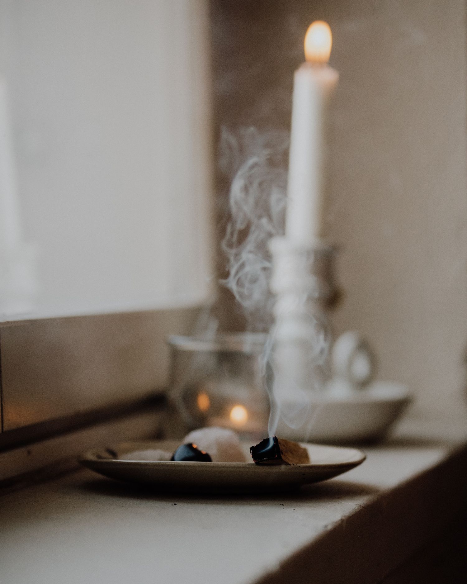 A lit white candle in a ceramic holder with wisps of smoke from burning incense on a dark plate nearby.