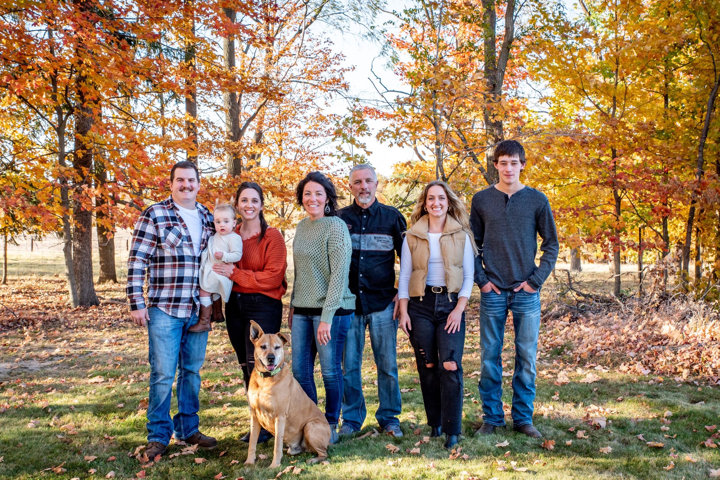 A group stands together outdoors with a dog against a backdrop of vibrant fall foliage in golden autumn light.