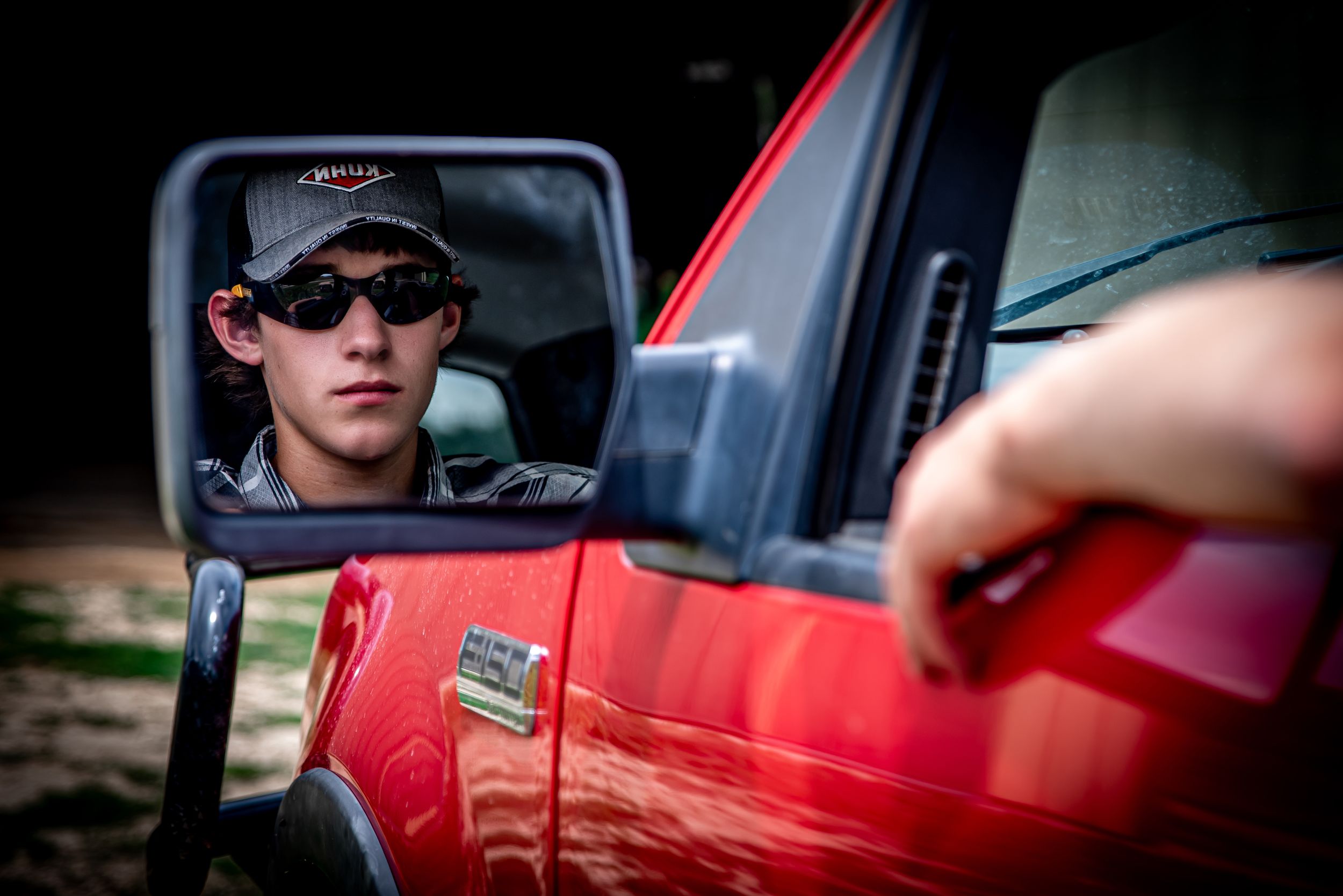 Side mirror reflection on a bright red vehicle showing sunglasses-wearing driver in the mirror.