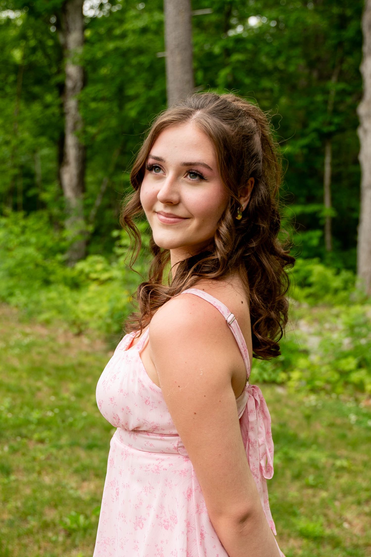 A woman in a pink dress stands in front of lush green trees while posing outdoors for a series of casual photos.