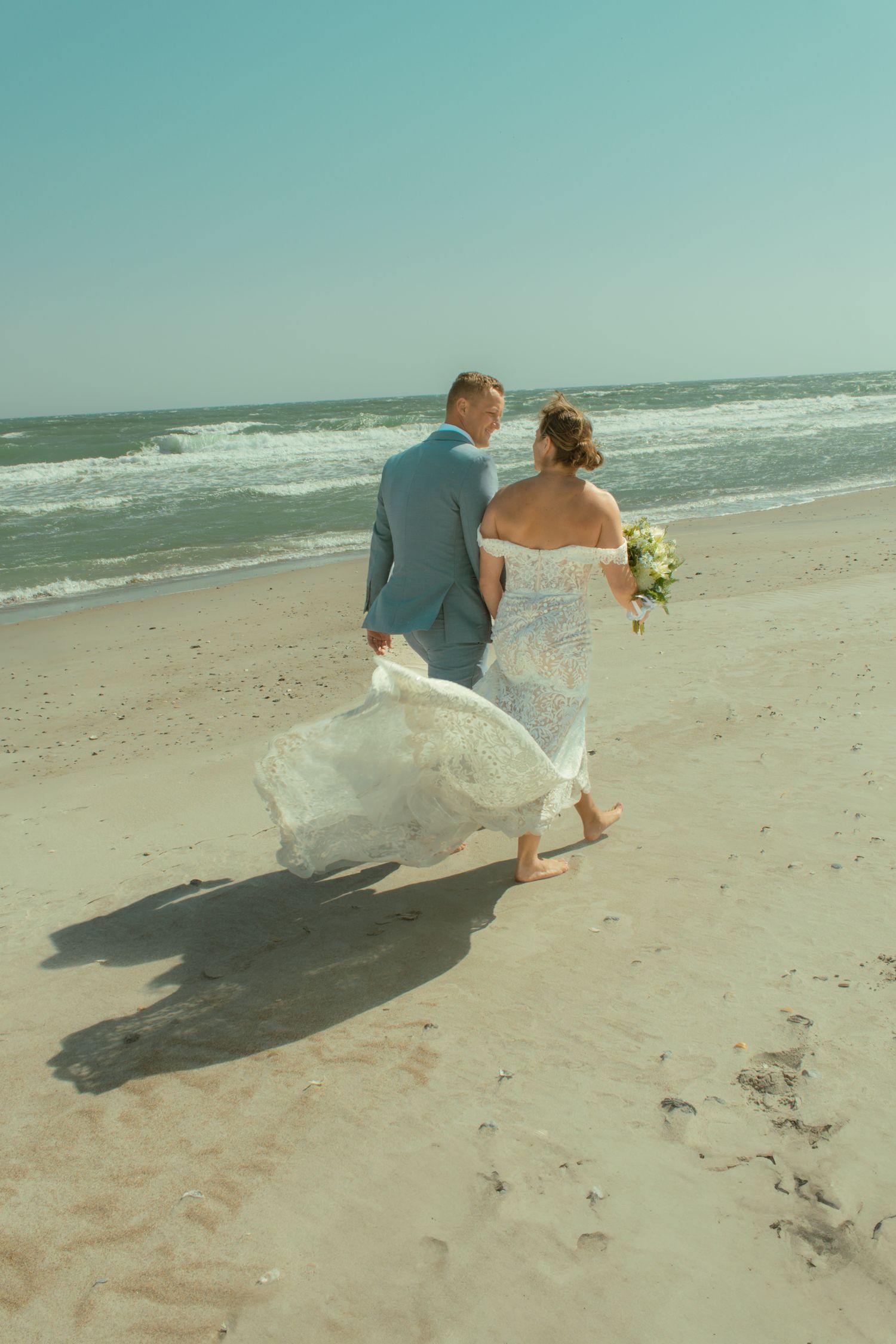 Newlyweds walking along beach shoreline with bride's dress flowing in the wind.