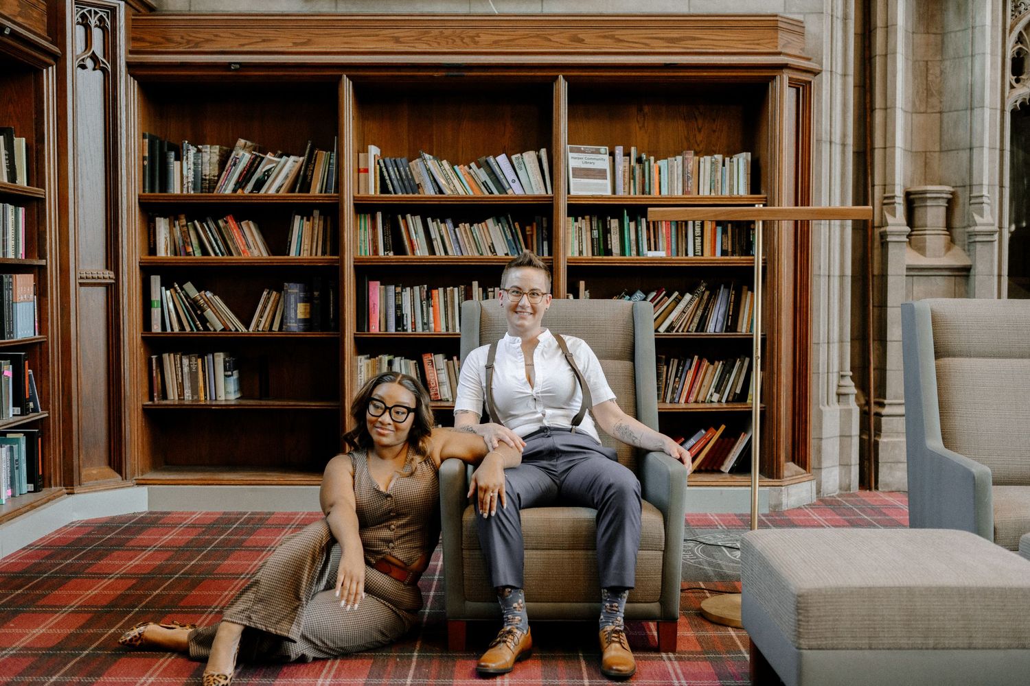 Modern couple sits together on floor in front of vintage wooden bookshelves in library setting.