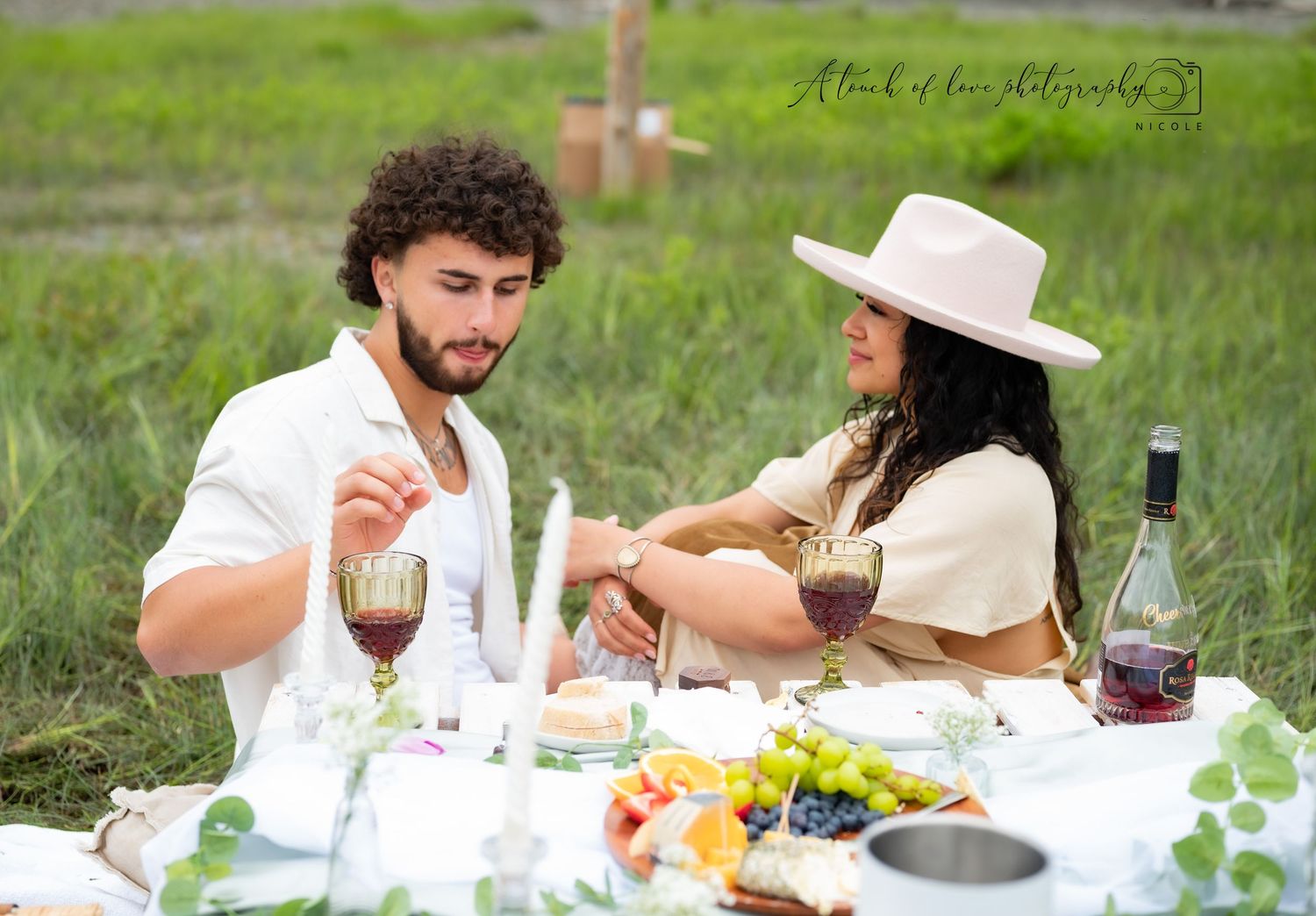 Couple enjoying a romantic outdoor picnic with wine and fruit on a blanket in a green field.