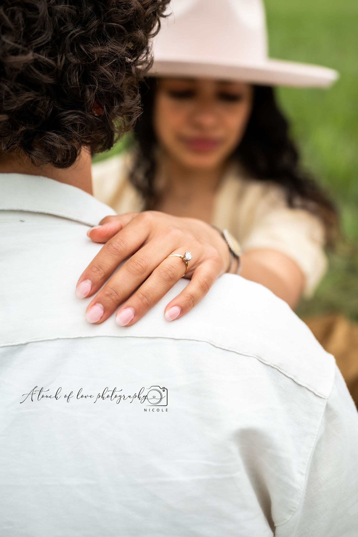 Woman in a wide-brim hat rests her hand on a man's shoulder, showing off her diamond engagement ring.