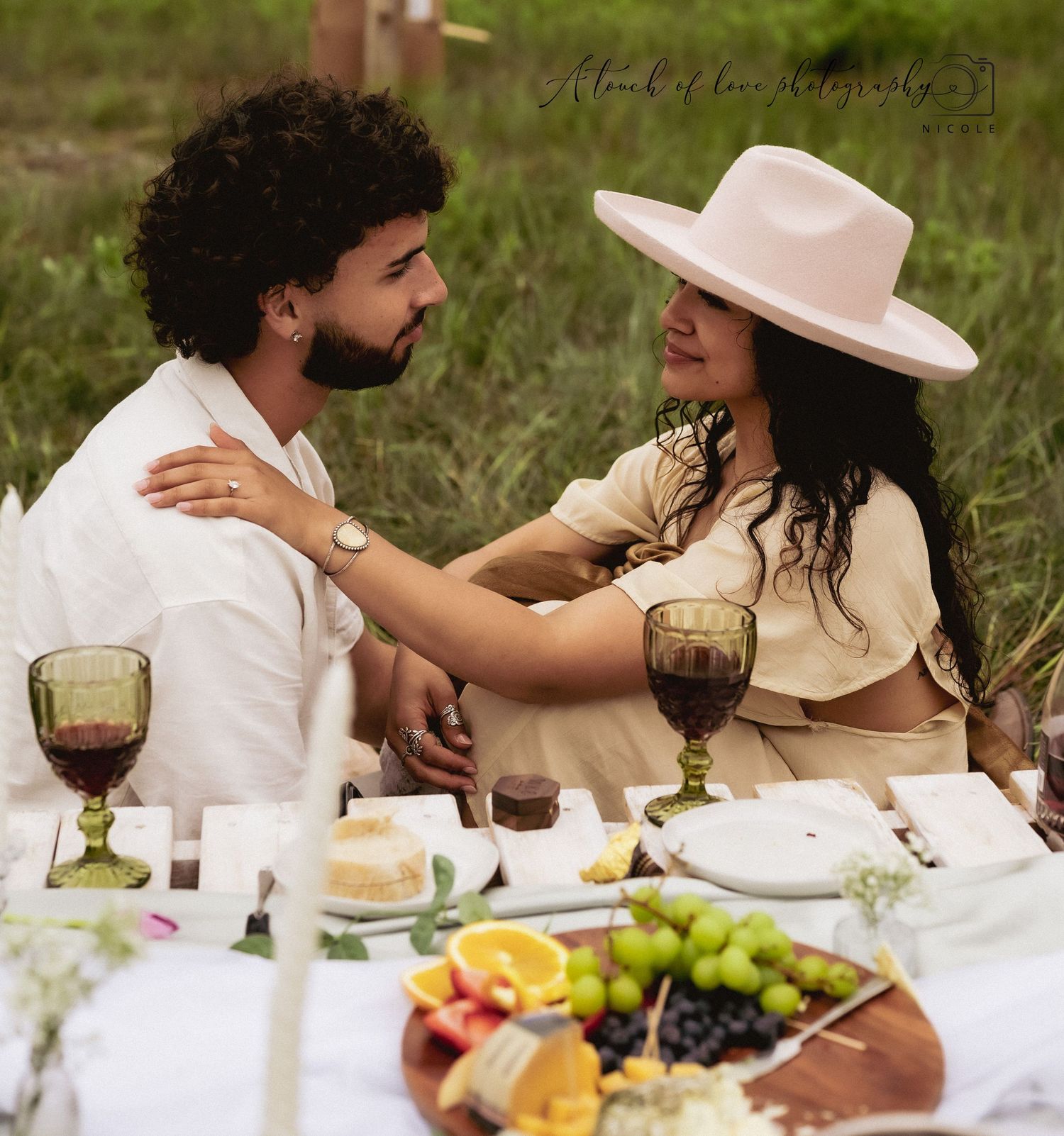 Couple sharing a romantic outdoor picnic with wine, fruit, and cheese, woman in pink hat gazing at man.