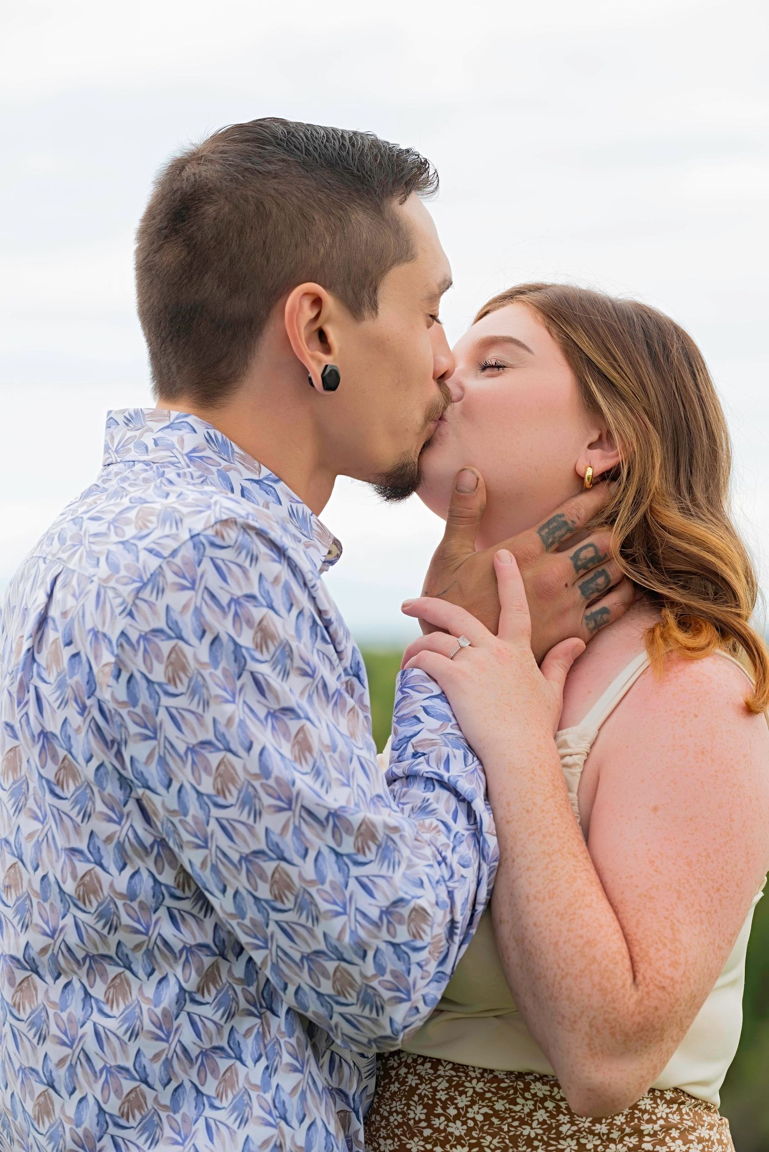 A couple in a romantic embrace sharing a tender kiss outdoors, with the person wearing a blue patterned shirt.
