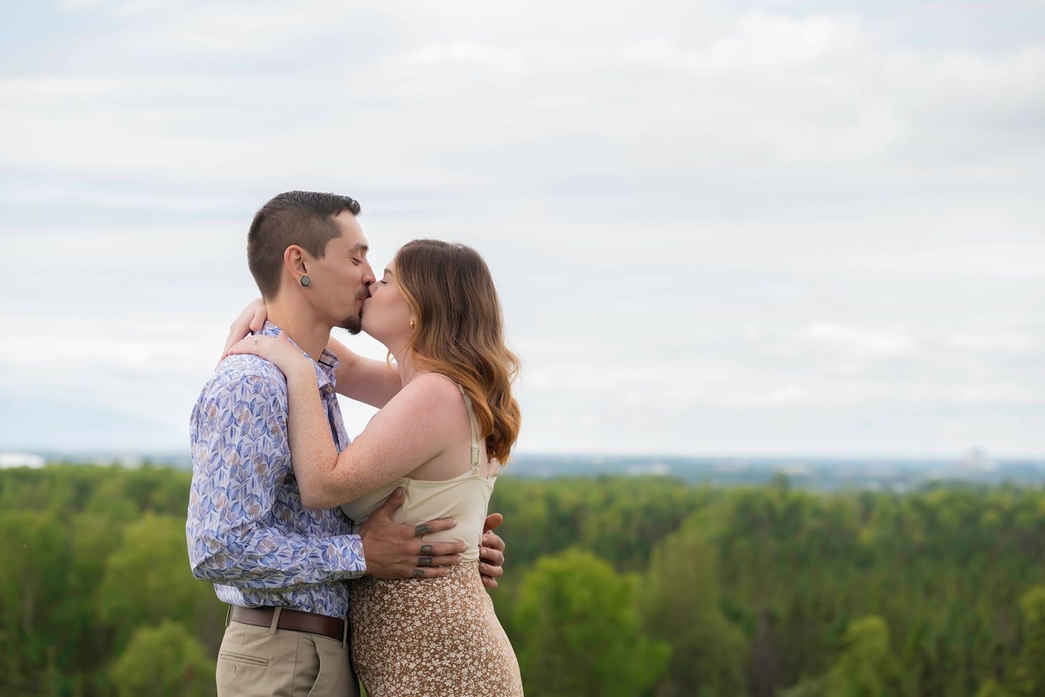 A couple embraces on a hilltop overlooking a scenic green valley landscape on an overcast afternoon.
