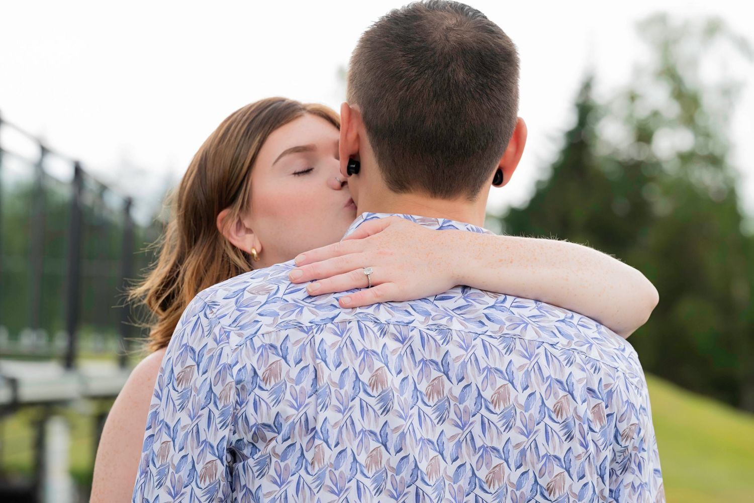 A couple shares an intimate embrace outdoors on a grassy hill while wearing coordinated blue and white outfits.