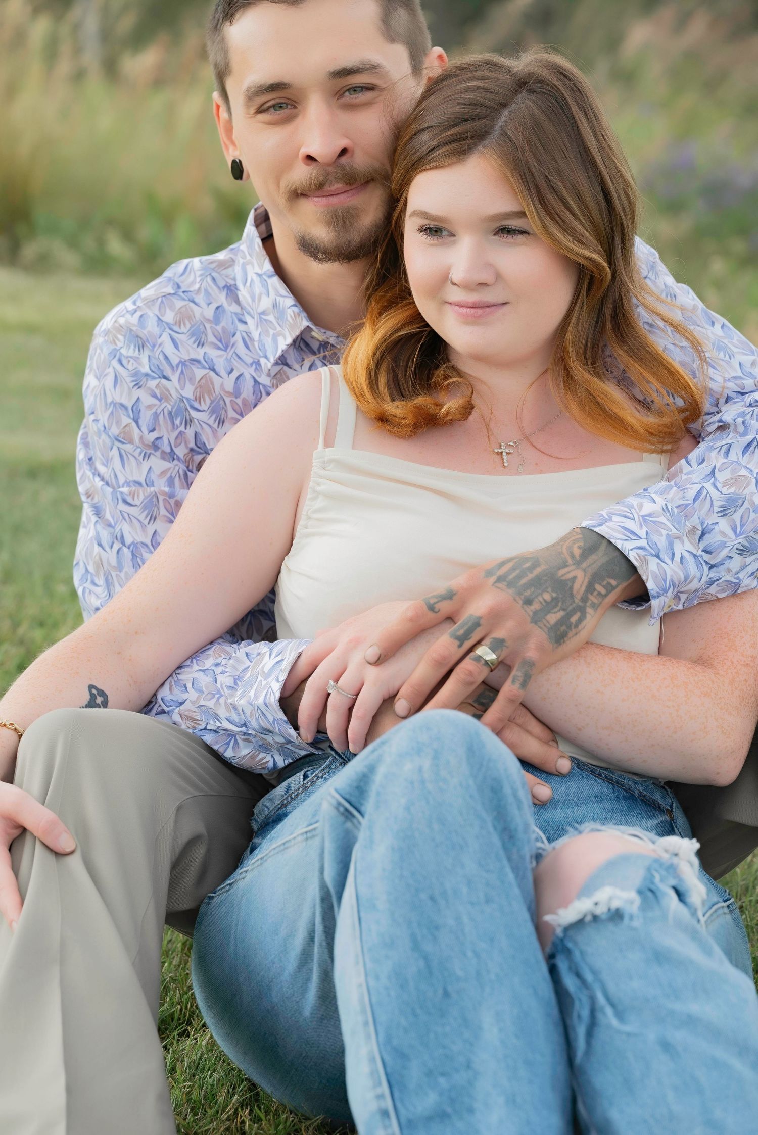 A couple sits together on grassy ground in casual attire during a spring outdoor photo session.