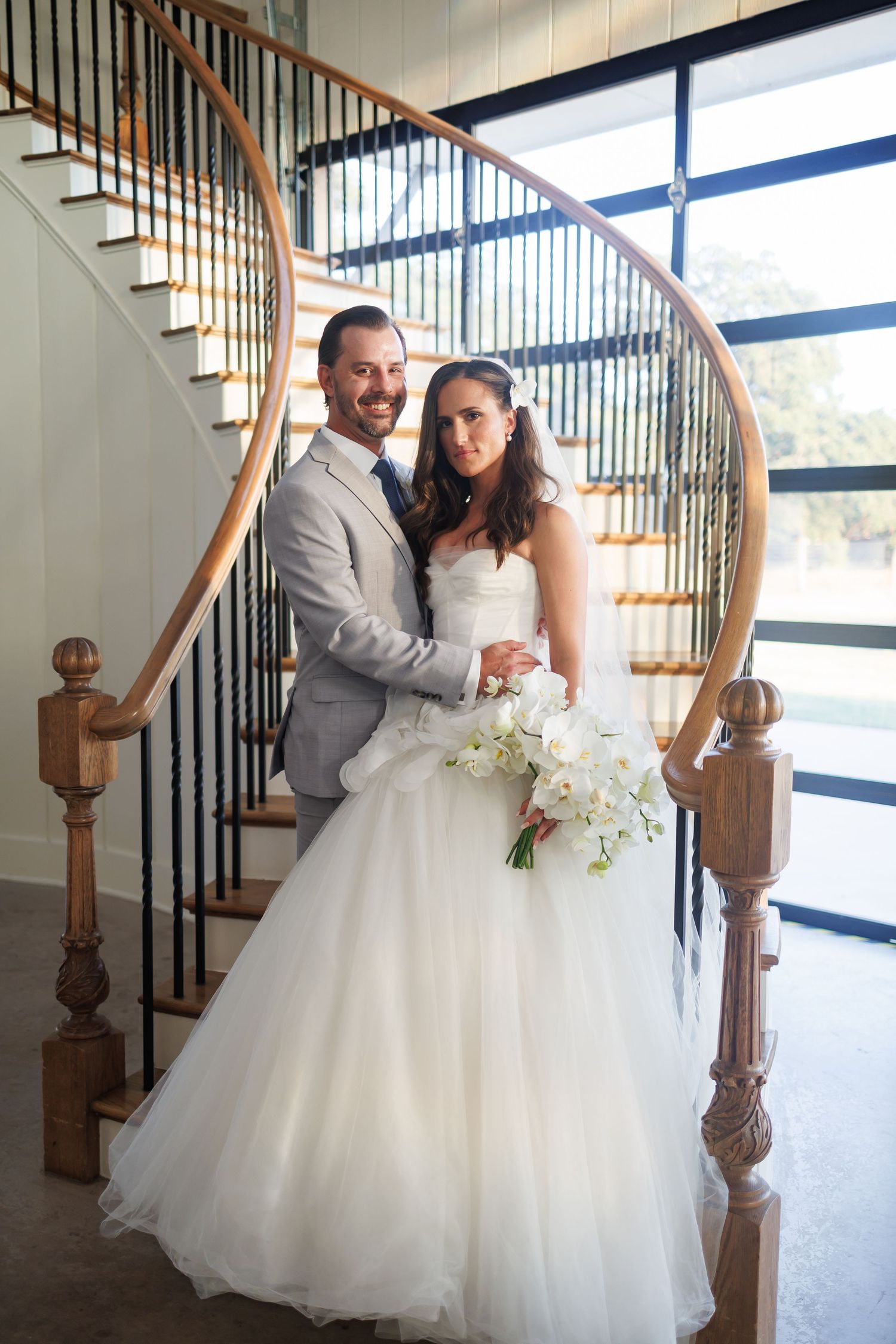 Couple poses on elegant curved staircase at Bella Cavalli with large windows, bride in flowing white gown holds white floral bouquet.