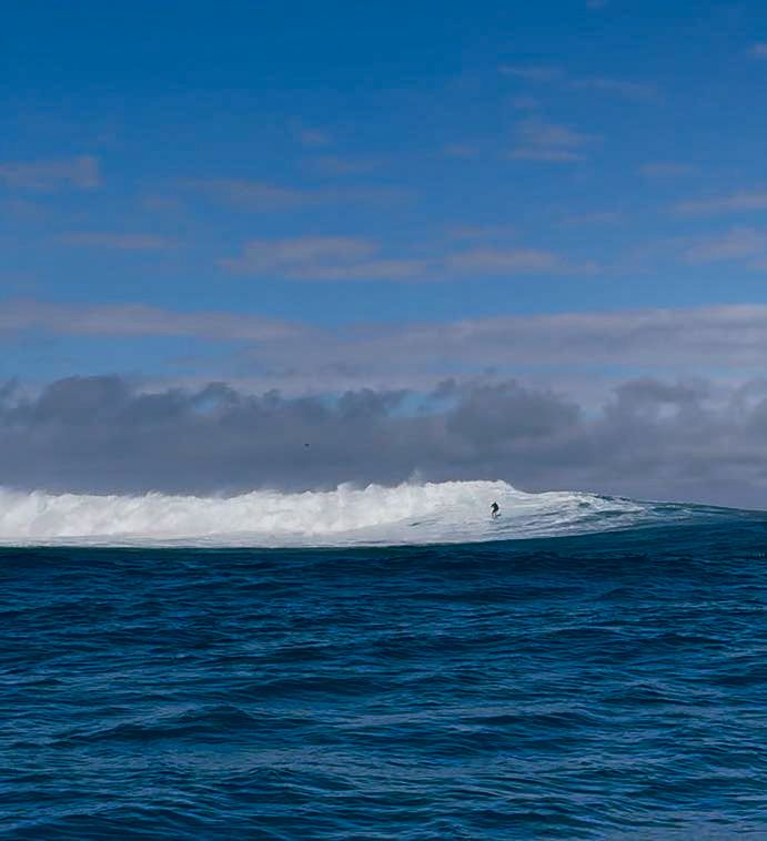 Vast blue ocean meets horizon under cloudy sky with distant iceberg visible.