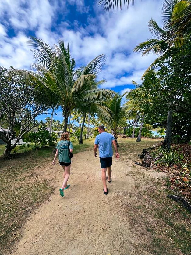 Two people walking down a dirt path lined with palm trees and tropical vegetation.