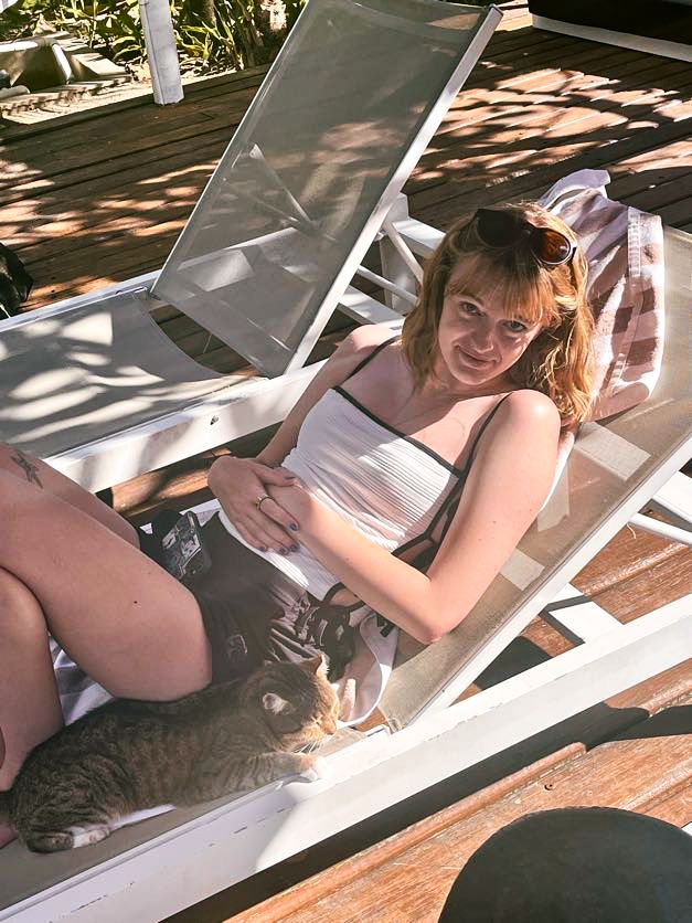 Relaxing on a white lounge chair with a cat on a sunny wooden deck.