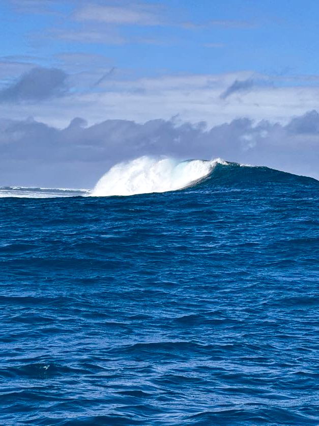 A large ocean wave breaks with white spray against deep blue waters under a cloudy sky.