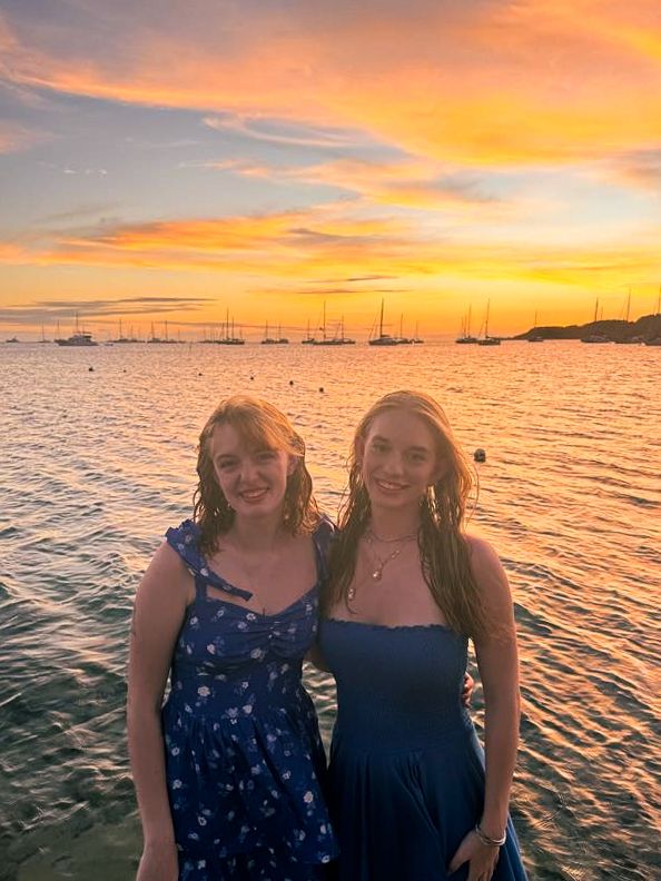 Two figures in blue dresses pose together at sunset with boats silhouetted in the harbor background.