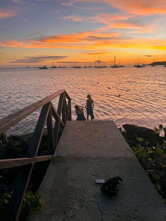 Silhouetted figures stand on a pier during a vibrant orange sunset over a harbor filled with boats.