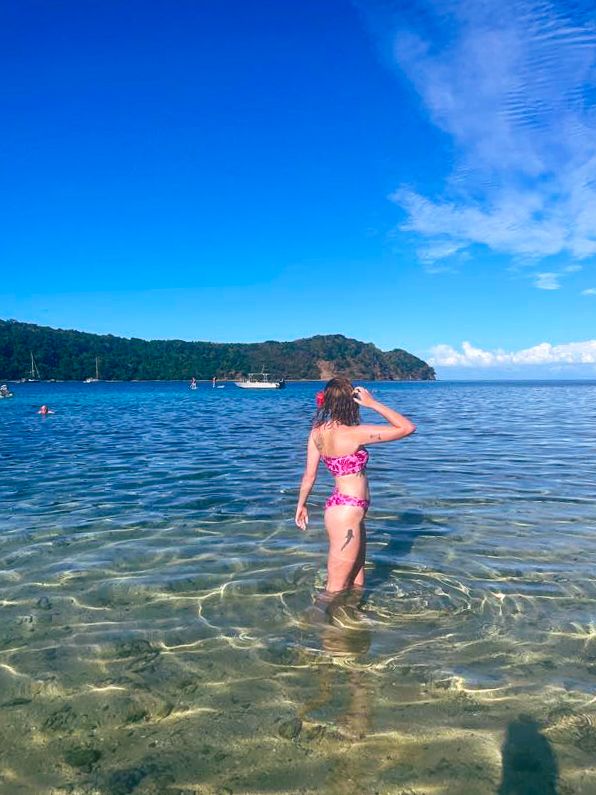 A person stands in shallow tropical waters looking out towards mountains and boats in the distance.
