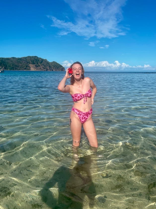 Person in pink floral bikini splashing and laughing in crystal clear tropical waters with mountains in background.