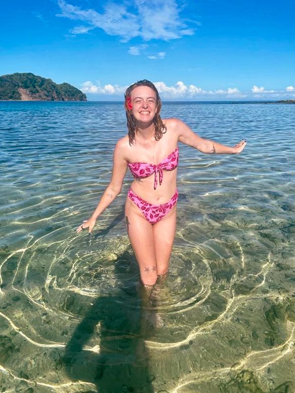 Person enjoying shallow turquoise waters with arms outstretched and mountains visible on the horizon.