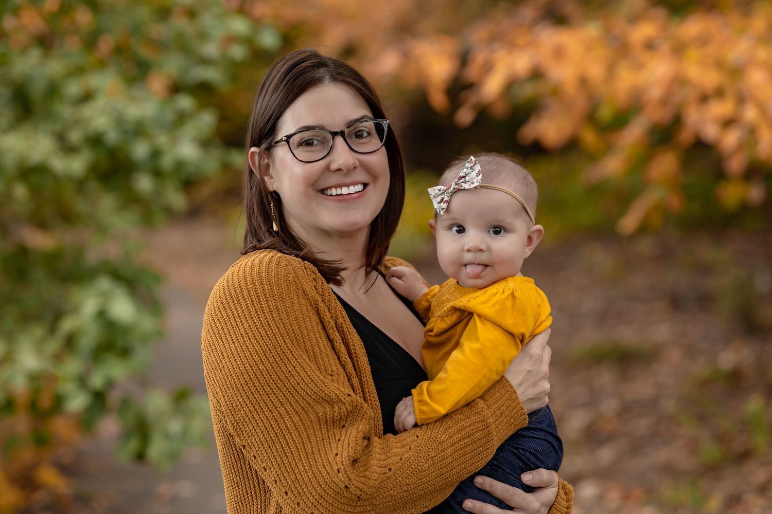 A parent wearing a brown cardigan holds a baby in a yellow sweater while standing amongst fall foliage. Style guide for fall mini session
