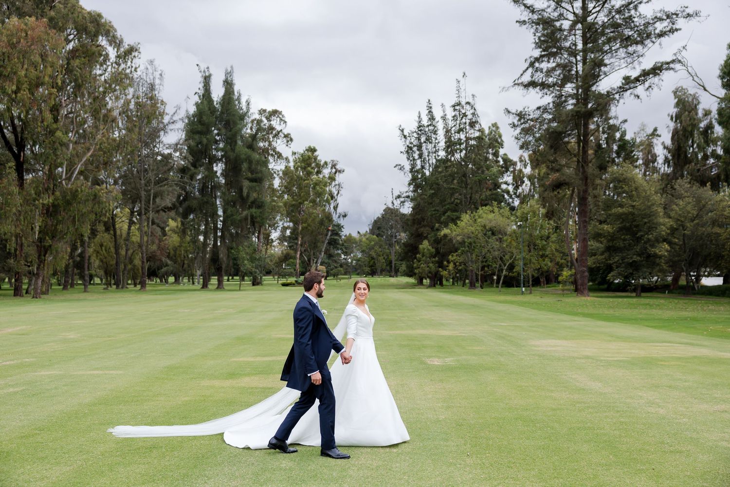 Wedding couple walks hand in hand across a scenic golf course with evergreen trees in background.