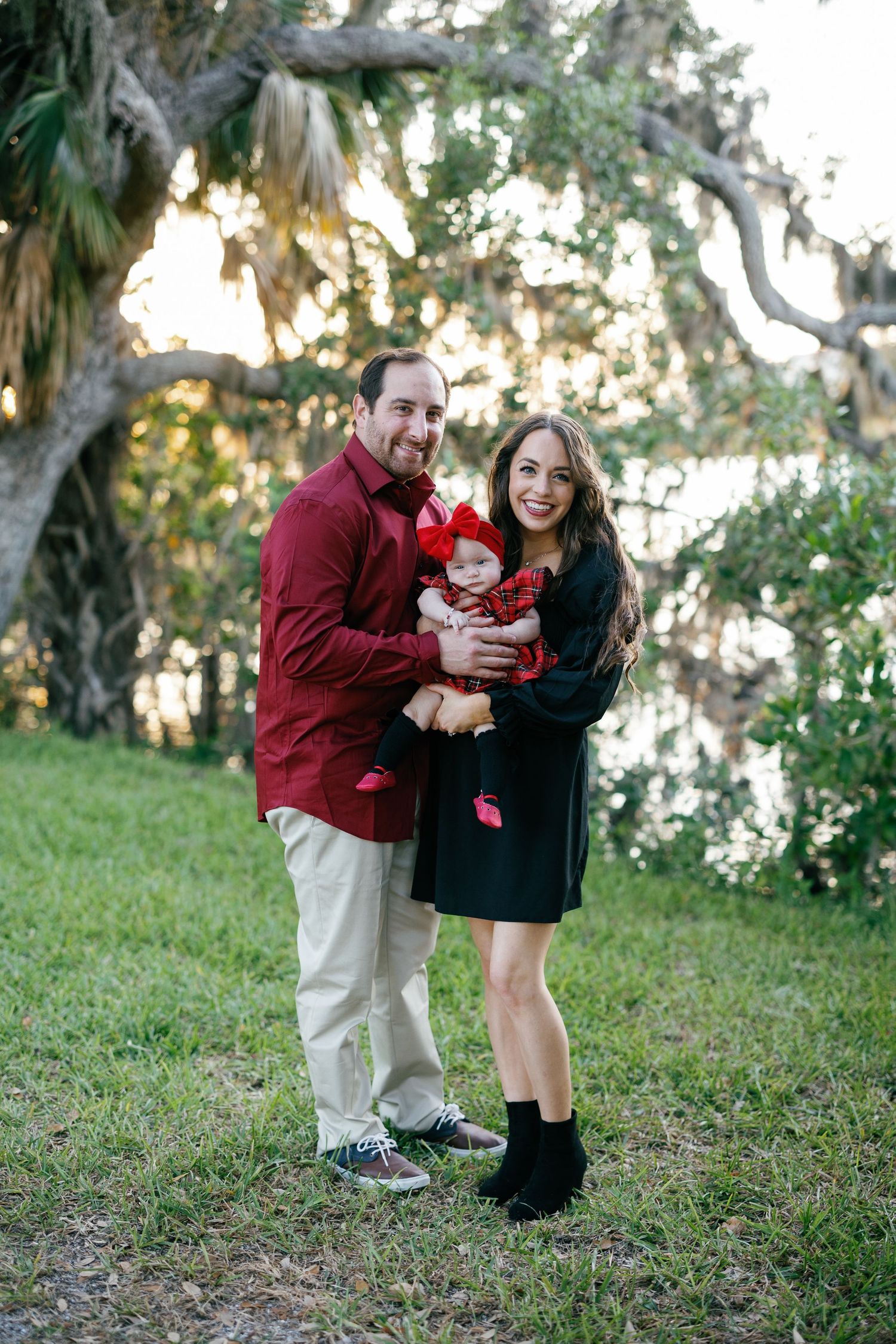 Family in matching red and black Christmas outfits pose together under a majestic oak tree at sunset.