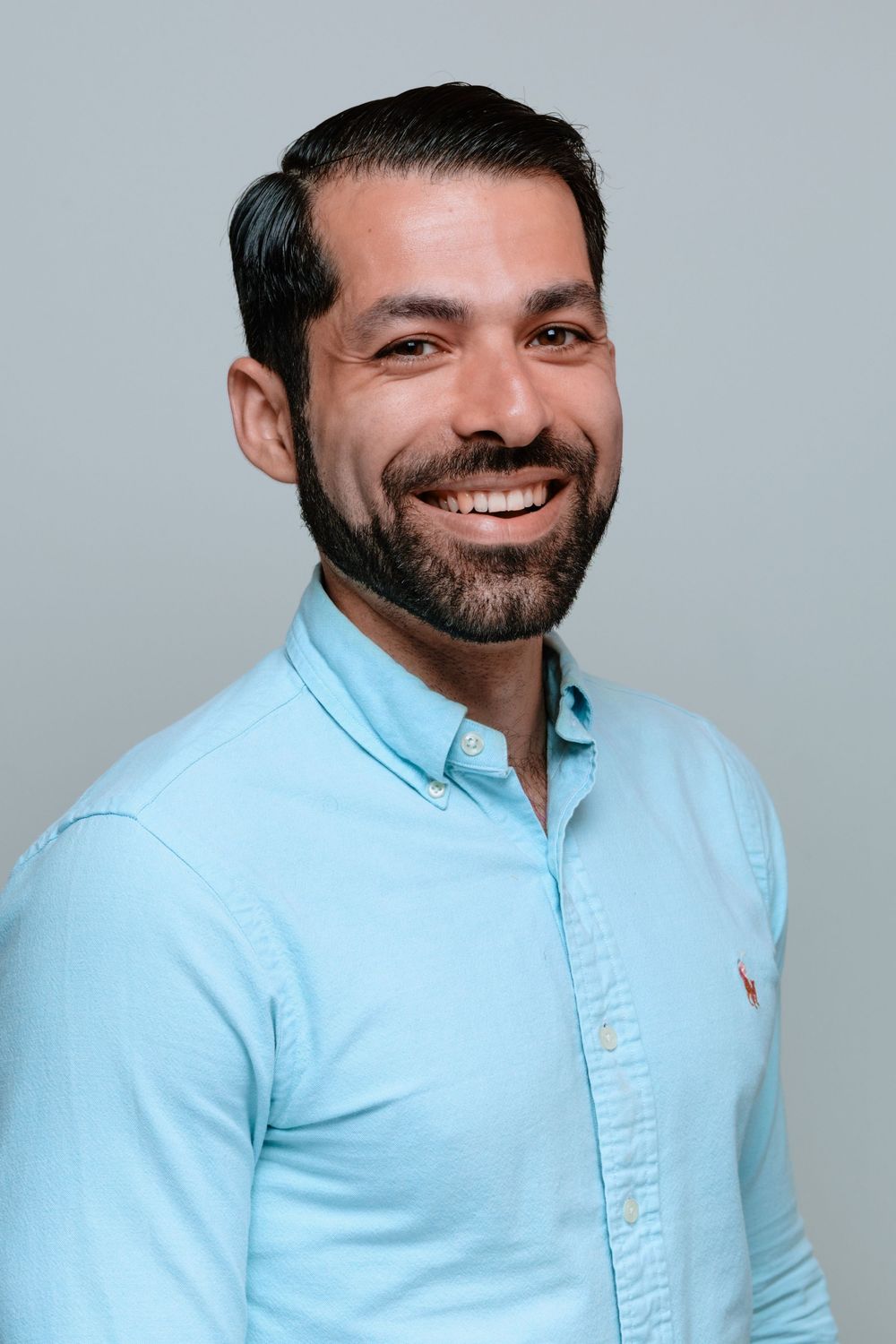 Professional headshot of someone wearing a light blue button-down shirt against a gray background, with a warm smile.
