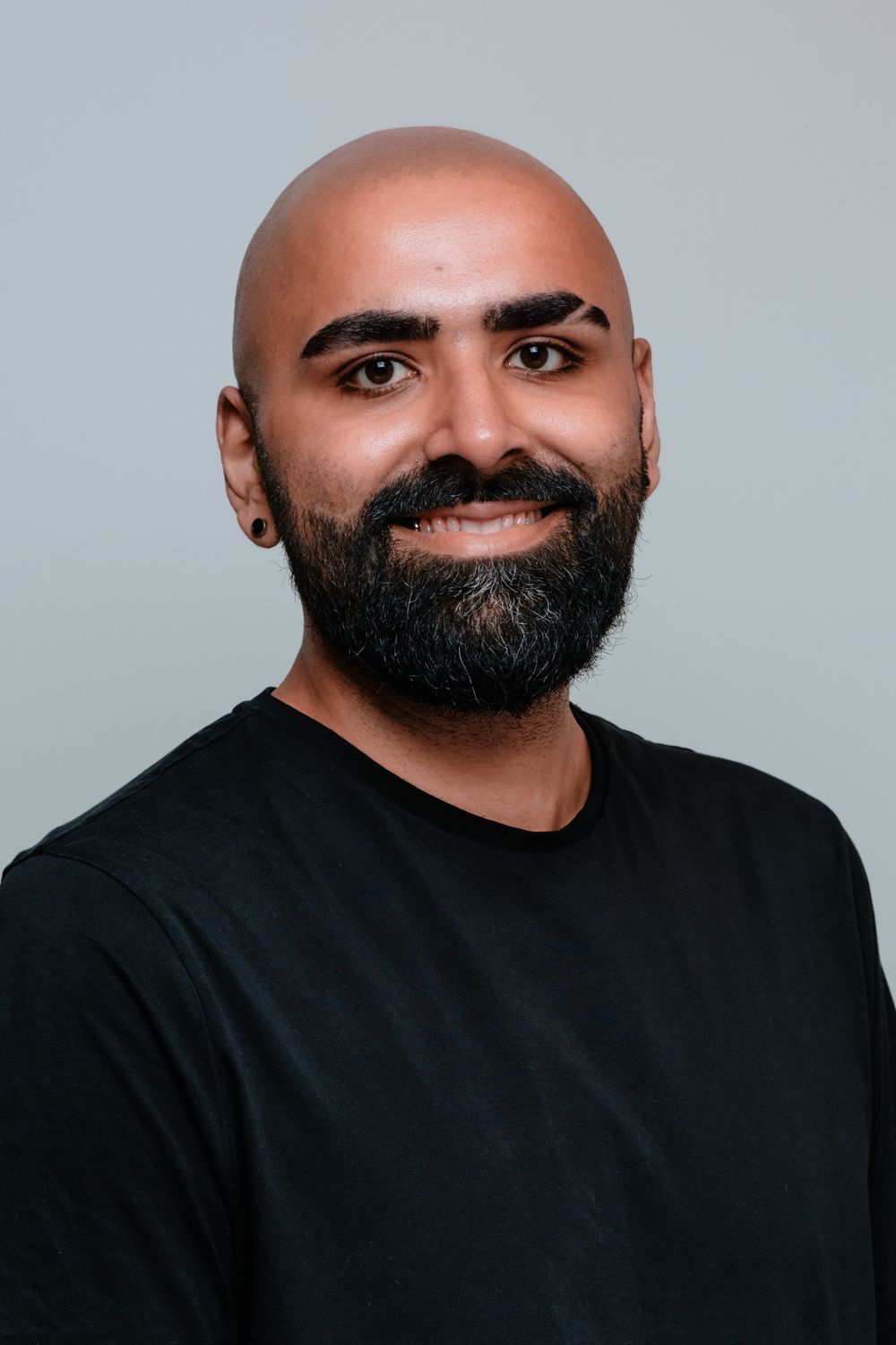 Portrait of bearded person in black shirt smiling against gray backdrop.