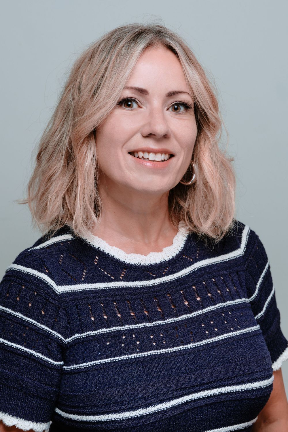 Professional headshot featuring a stylish navy striped shirt and warm smile against a neutral backdrop.