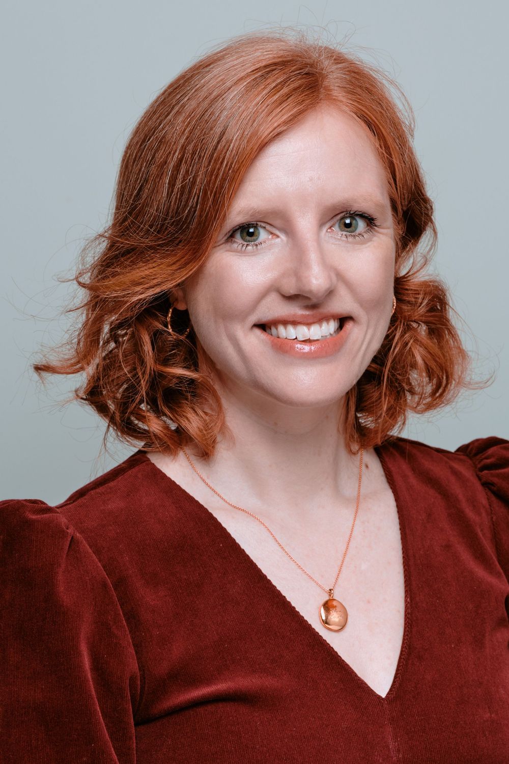 A cheerful redhead with curly hair poses against a neutral backdrop in a burgundy top.