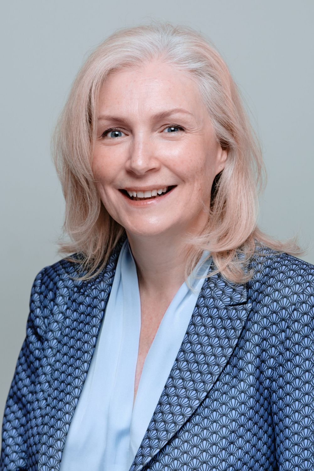 Professional headshot series showing joyful expressions in a blue patterned blazer and light blouse against gray background.
