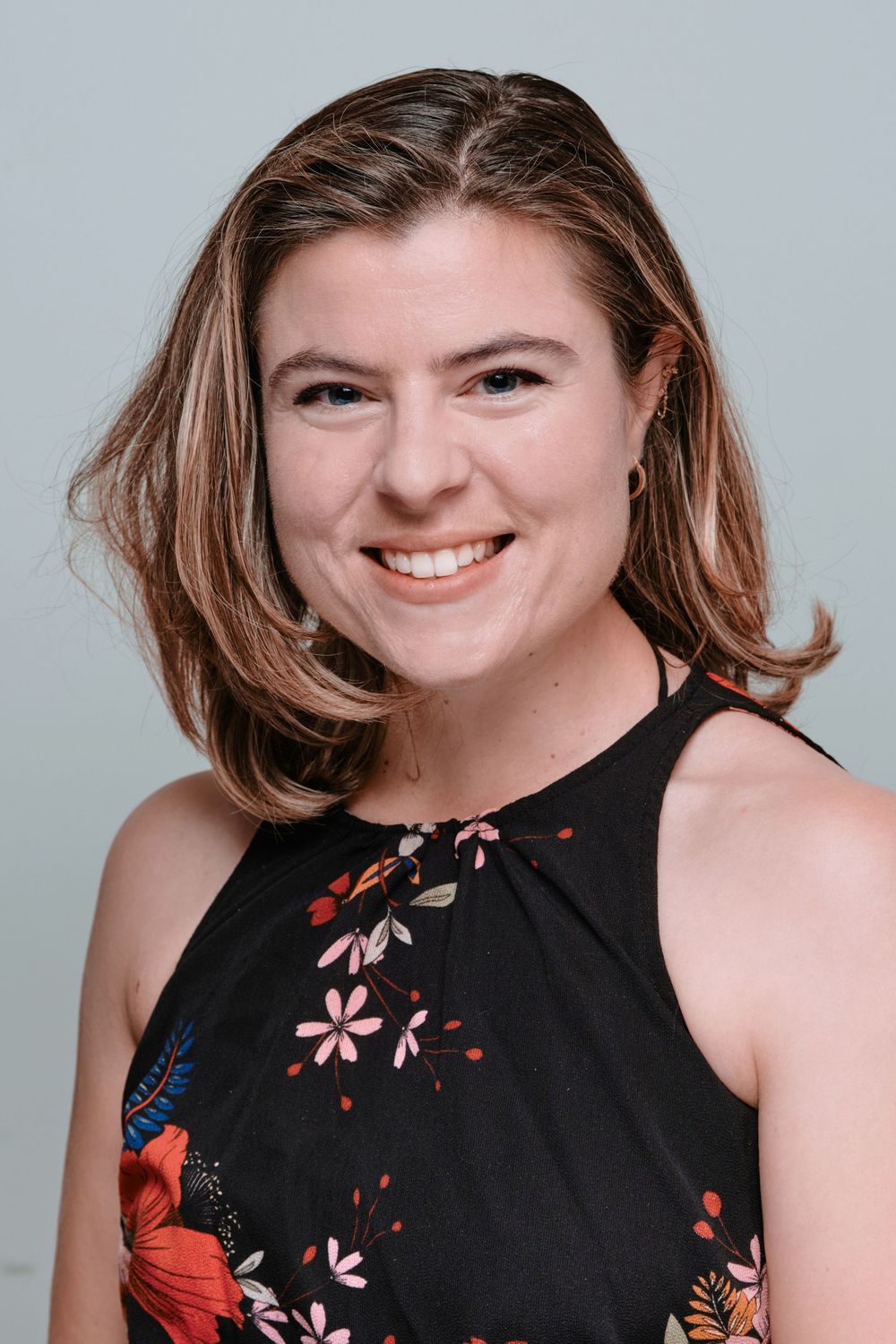 Professional headshot of someone in a black floral sleeveless top smiling warmly against a light gray background.