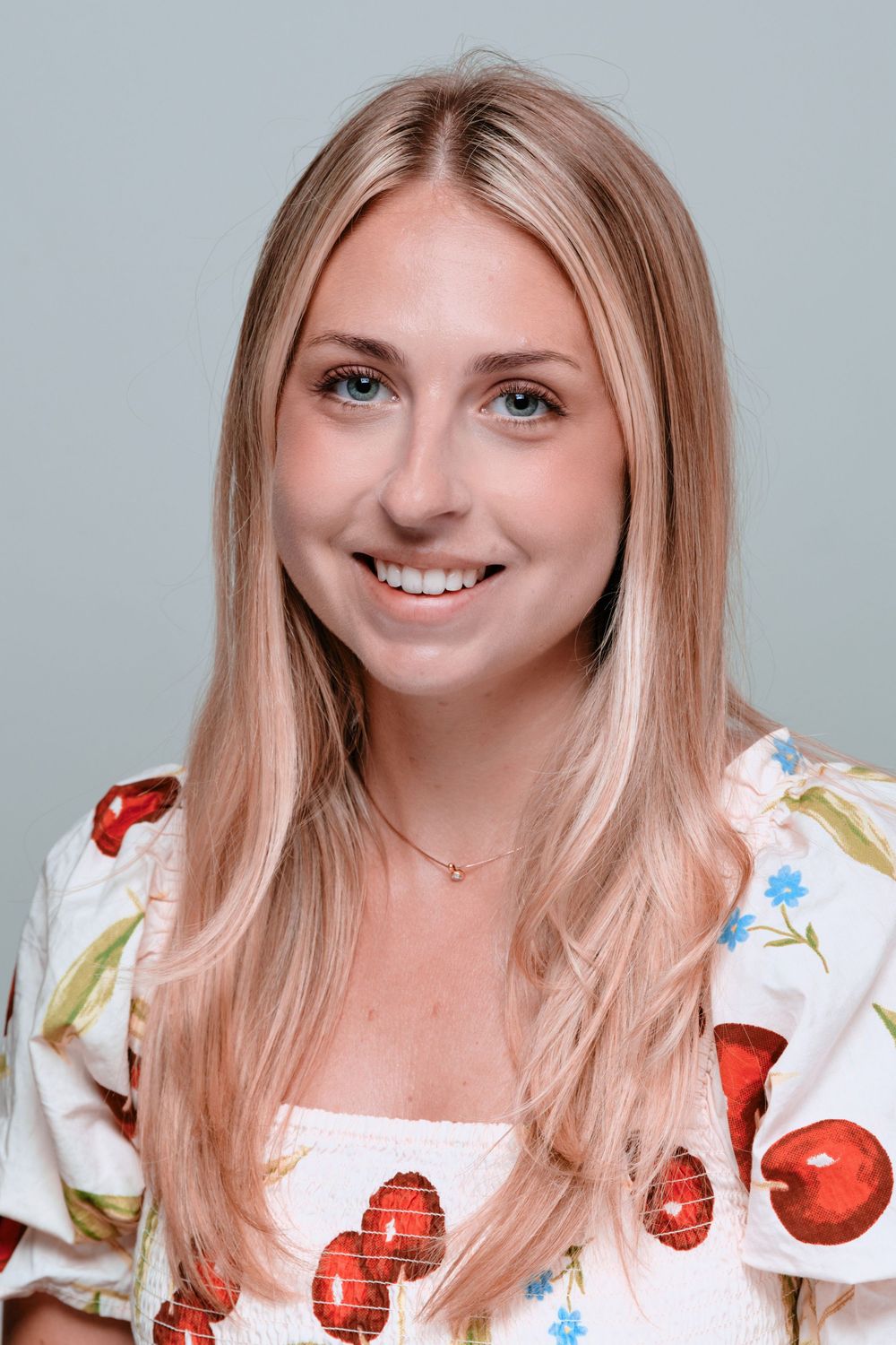 Smiling person with long blonde hair wearing a floral print dress with red flowers against a light gray background.
