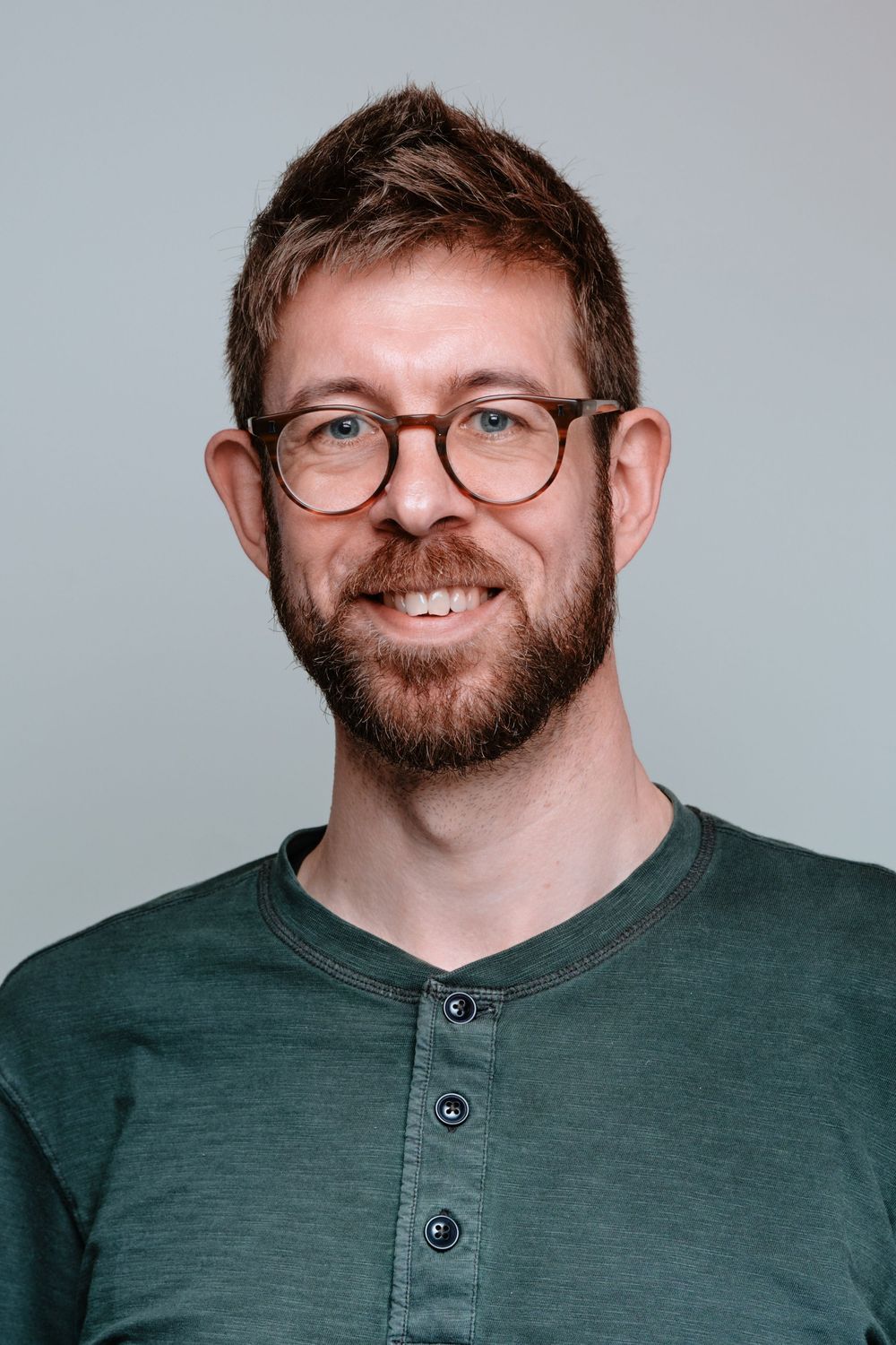 Professional portrait of a person with glasses and a beard wearing a green henley shirt against a gray background.
