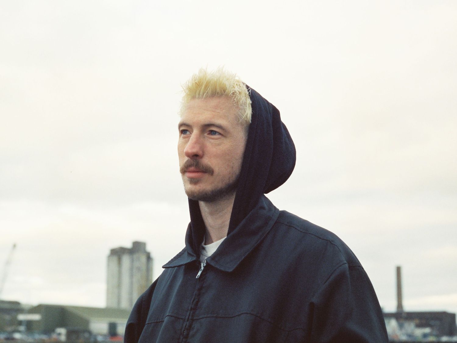 A person with bright blonde hair and dark hoodie sits thoughtfully against a cloudy outdoor backdrop.