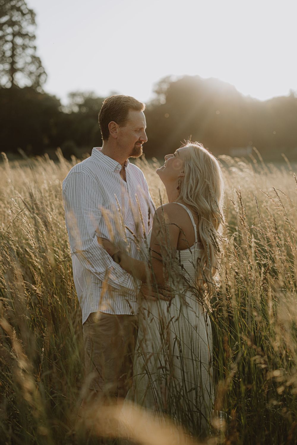 Romantic couple embraces in golden sunlit field of tall grass during sunset photoshoot.