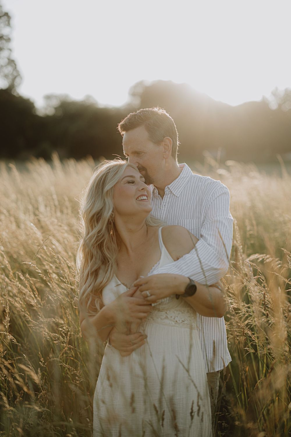 A couple laughing together in a sunlit field of tall grass during golden hour.