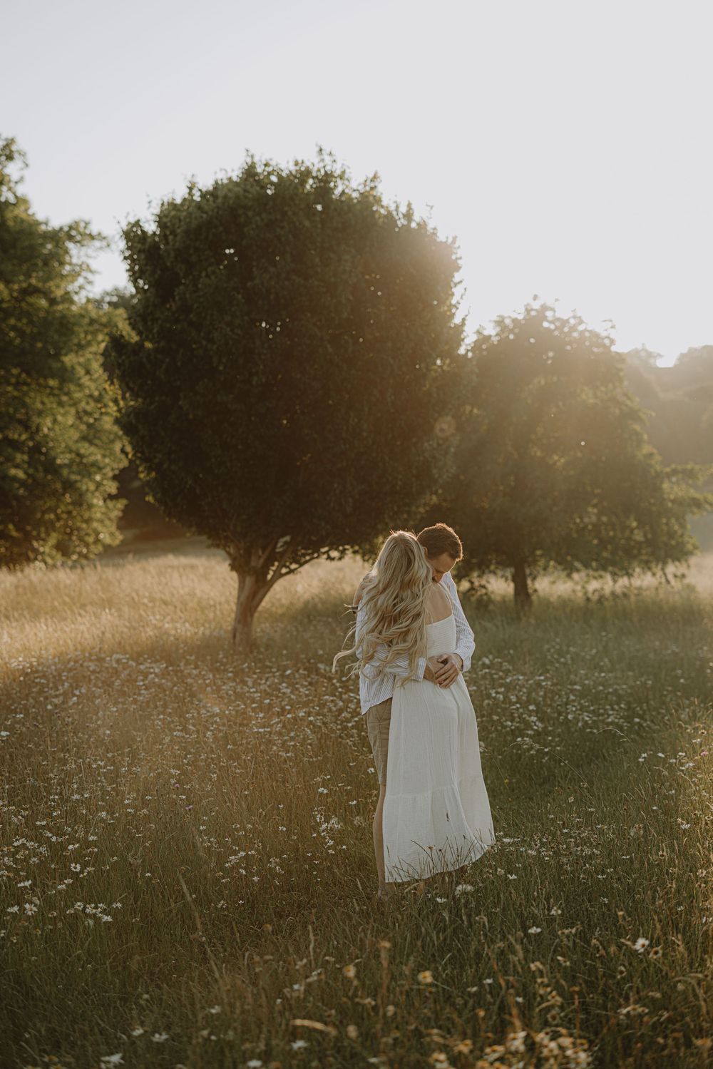 Couple shares intimate moment in sunlit field with trees in background during golden hour.